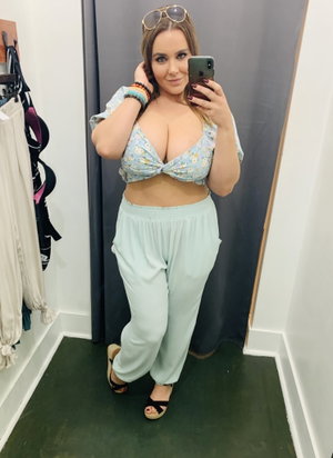 Woman in floral top and light pants takes mirror selfie in fitting room.