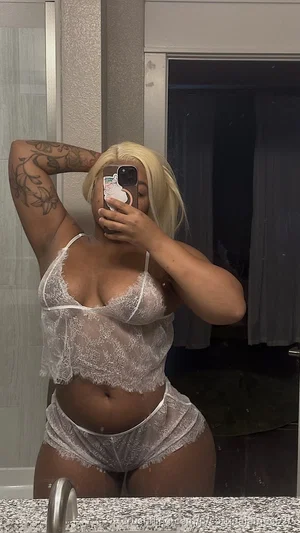 Woman with blonde hair and tattoos wears white lace lingerie, taking mirror selfie in bathroom.