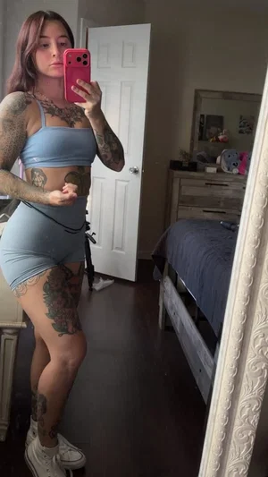 Woman with tattoos in blue crop top and shorts takes mirror selfie in bedroom.