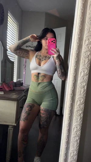 Woman with tattoos in white crop top and green shorts taking mirror selfie in bedroom.