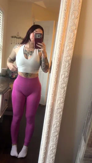 Woman with tattoos in white crop top and purple leggings takes mirror selfie in bedroom.
