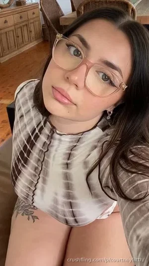 Woman with dark hair and glasses in a tie-dye top, sitting indoors with a tattoo on her side.
