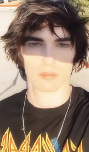 Man with dark messy hair in black t-shirt and chain outdoors.