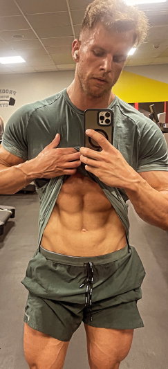 Fit man lifting shirt to show abs in gym mirror selfie.