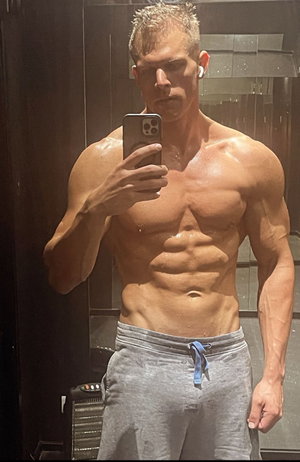 Muscular man taking mirror selfie shirtless in grey sweatpants.