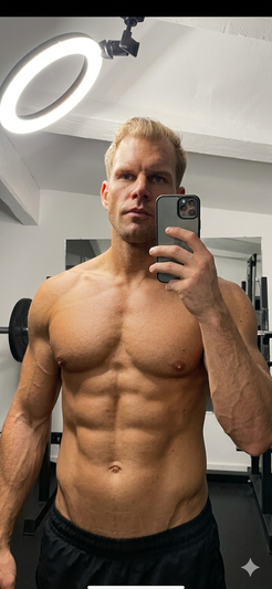 Shirtless muscular man with short blonde hair takes mirror selfie in gym, wearing black shorts.