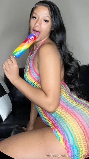Woman in colorful mesh dress holding rainbow popsicle, sitting on couch indoors.