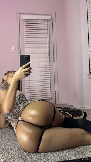 Woman in black thong and lace top lying on carpet in pink room, taking mirror selfie.