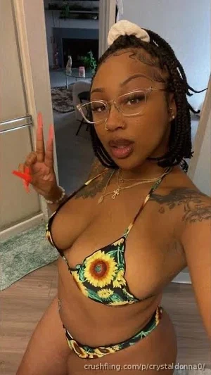 Woman with braided hair and glasses in floral bikini, making peace sign indoors.