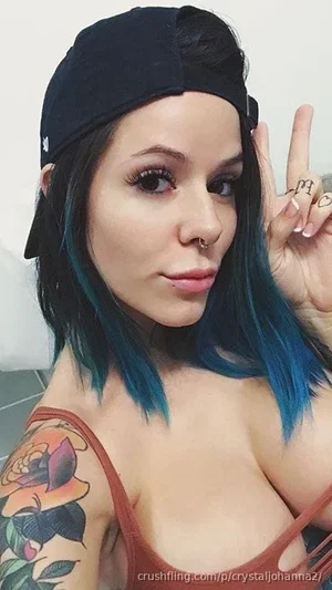 Woman with blue-tipped hair, nose ring, and tattoo, wearing a red top and black cap, posing indoors.