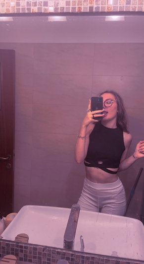 Brunette woman with glasses taking mirror selfie in bathroom wearing black crop top.