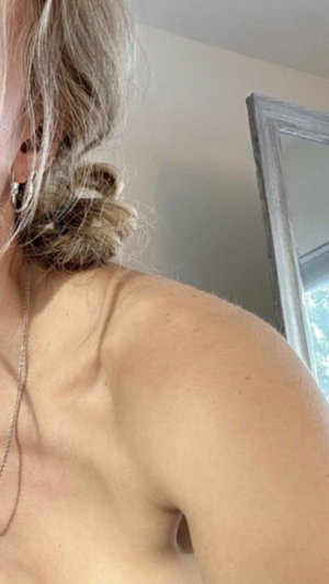 Woman with blonde hair in updo, bare shoulders, wearing necklace, mirror visible.