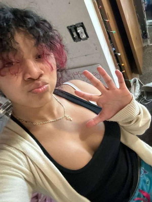 Woman with curly pink-tipped hair in black top taking indoor selfie.