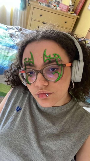Woman with curly hair, glasses, and green face art, wearing headphones and a gray tank top in a