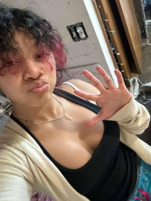 Woman with curly hair and pink highlights wearing black top and cream cardigan, waving in indoor