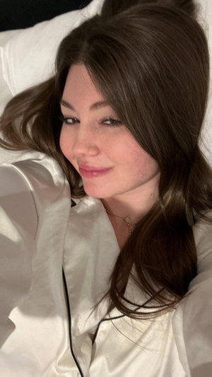 Woman with long brown hair in white satin pajamas lying in bed.