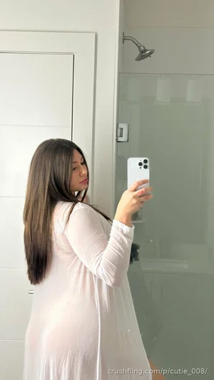 Woman with long dark hair in a sheer white dress taking a mirror selfie in a bathroom.
