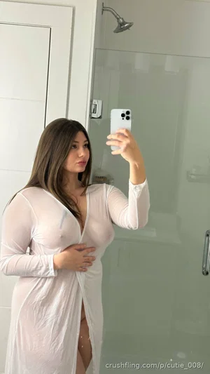 Woman in sheer white dress taking mirror selfie in bathroom, holding phone.