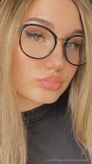 Blonde woman with glasses and makeup, wearing a black top, close-up selfie.