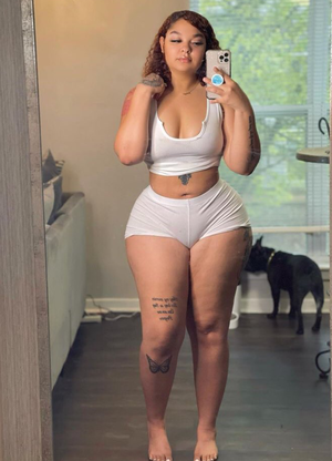 Woman in white crop top and shorts takes mirror selfie in bedroom, tattoos visible on arms and legs.