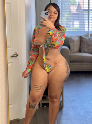 Woman in floral top and matching thong, tattoos on legs and hip, taking mirror selfie in living