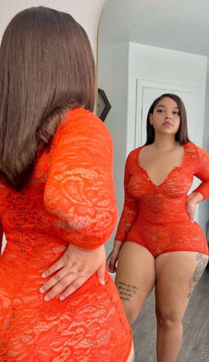Woman in red lace bodysuit posing before mirror indoors.