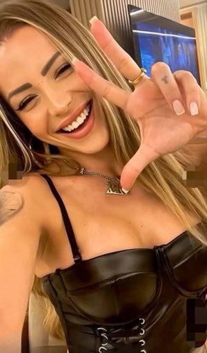 Woman with blonde hair in black leather corset top, smiling and making peace sign indoors.