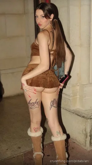 Woman in brown velvet outfit with thigh tattoos, standing indoors near stone wall.