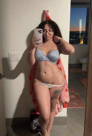 Woman in light blue bra and white thong takes mirror selfie in bathroom, curly hair, glasses,