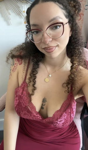 Woman with curly hair and glasses wears a red lace top, sitting indoors.