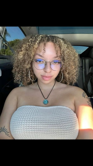 Woman with curly hair and glasses in a white strapless top, sitting in a car.