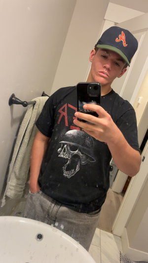 Young man in black skull t-shirt and cap takes mirror selfie in bathroom.