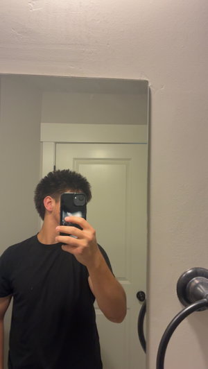 Man in black t-shirt taking mirror selfie in bathroom.