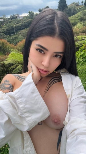 Woman with dark hair and tattoos in open white shirt outdoors.