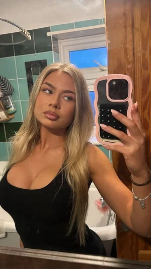 Woman with long blonde hair in black top taking mirror selfie in bathroom.