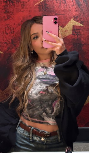 Woman with wavy hair in crop top, open hoodie, jeans, red wall selfie.