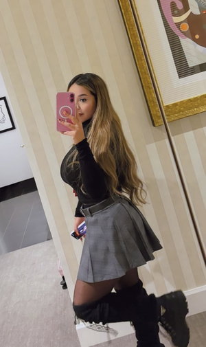 Curvy blonde woman in black top and plaid skirt taking mirror selfie.