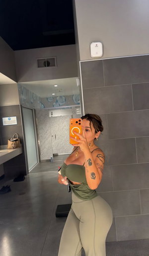 Woman with tattoos in green activewear taking mirror selfie in bathroom.