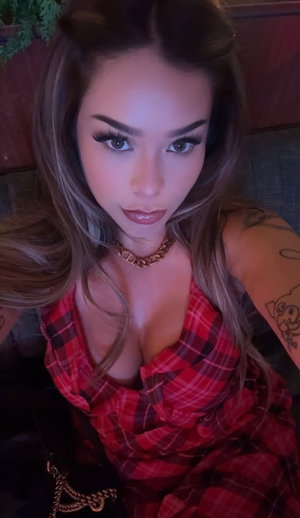 Woman with long hair and tattoos wears red plaid top, indoors.