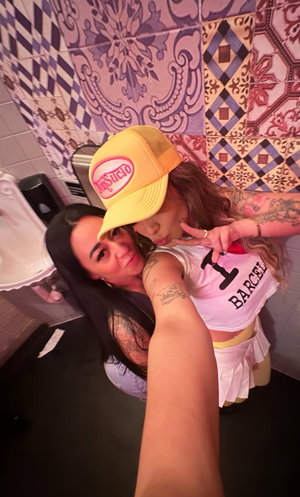 Two women with tattoos pose together in a tiled bathroom, one wearing a yellow cap and pink crop