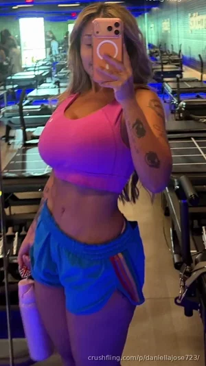 Woman in pink sports bra and blue shorts taking mirror selfie in gym.