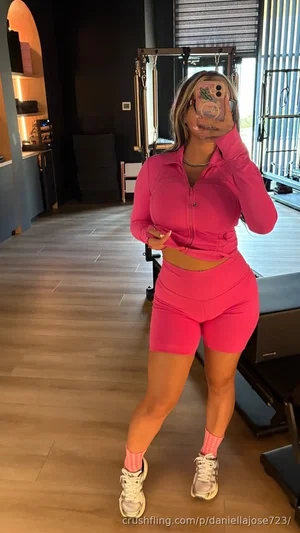 Woman in pink crop top and shorts takes mirror selfie in gym setting.