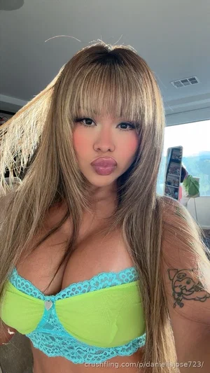 Woman with long blonde hair wearing green and blue lace bra, posing indoors.