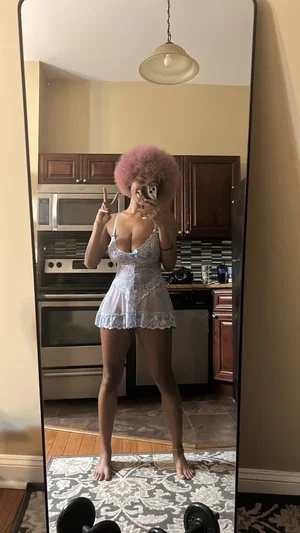 Woman with pink afro in white lace lingerie takes mirror selfie in kitchen.