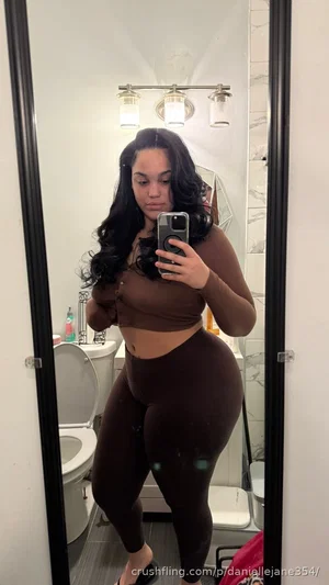 Woman with long dark hair in brown crop top and leggings, taking mirror selfie in bathroom.