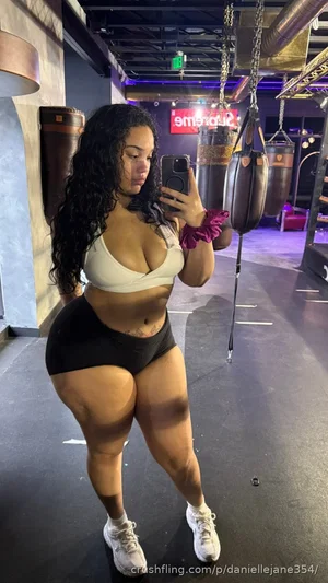Woman with curly hair in white sports bra and black shorts taking mirror selfie in gym.