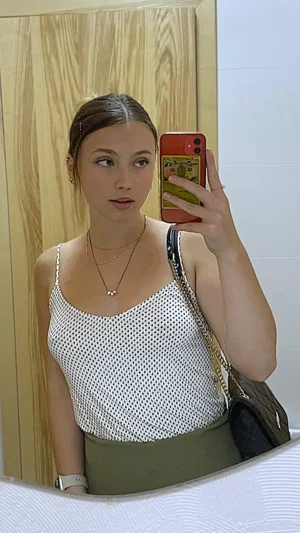 Woman in white polka dot top and green skirt takes mirror selfie in bathroom.
