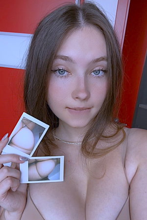 Brunette in low-cut beige top holds breast and butt Polaroids indoors.