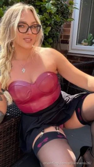 Blonde woman in red bustier and black stockings sitting outdoors on patio furniture.