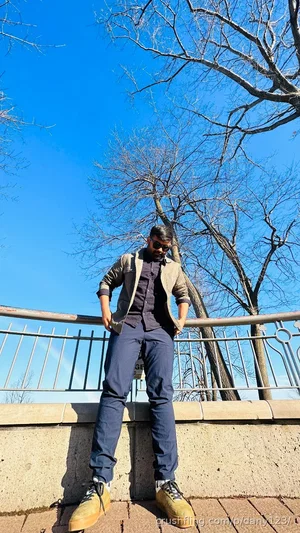 Man in casual outfit standing outdoors near a railing with trees and blue sky.